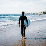 A surfer in a wetsuit carrying a foam board to the water at a beach near Lisbon